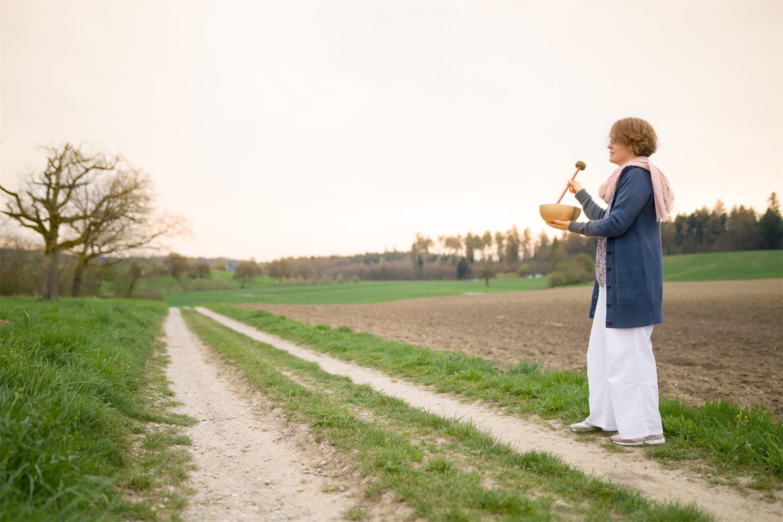 Eine Frau mit Klangschale steht in der Abenddämmerung auf einem Feldweg.