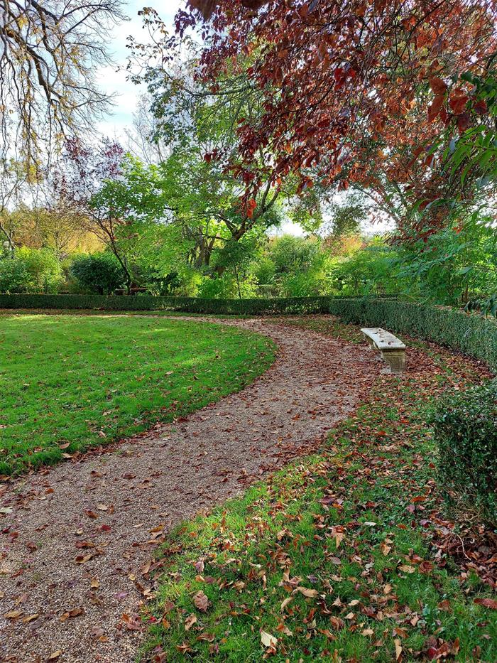 Ein Kiesweg führt durch einen herbstlichen Garten, am Rand steht eine Steinbank.